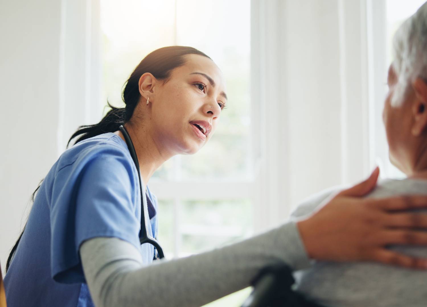 A nurse in blue scrubs comforting an elderly patient by placing a hand on their shoulder. 